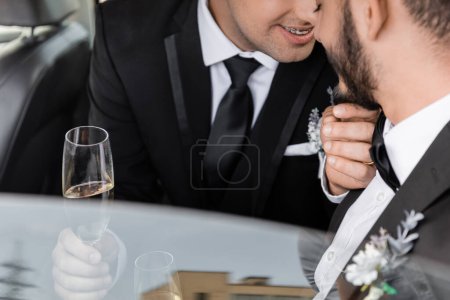 Cropped view of positive gay groom with braces in classic suit touching chin of bearded boyfriend and holding champagne after wedding celebration in car 