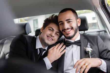 Joyeux marié homosexuel avec bretelles en costume étreignant copain barbu et regardant la caméra après la célébration du mariage tout en allant en lune de miel 