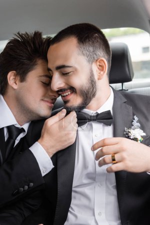 Young brunette gay groom in elegant formal wear touching chin of bearded boyfriend with closed eyes after wedding celebration while going on honeymoon  magic mug #654380392