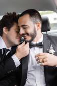 Young brunette gay groom in elegant formal wear touching chin of bearded boyfriend with closed eyes after wedding celebration while going on honeymoon  Poster #654380392