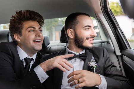 Cheerful gay groom in braces and elegant suit pointing with finger near bearded boyfriend and looking together through car window after wedding celebration while going on honeymoon 