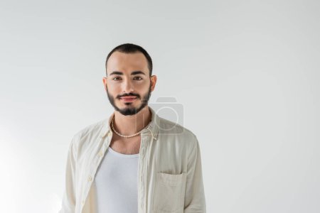 Retrato de joven hombre homosexual de pelo corto y barbudo en collar de perlas y ropa casual mirando a la cámara mientras está de pie aislado en gris  