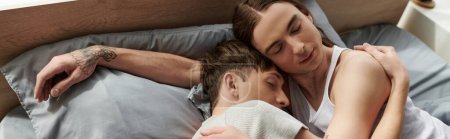 Téléchargez les photos : Vue grand angle de tatoué et jeune couple gay en pyjama serrant doucement l'un l'autre tout en dormant ensemble sur un lit confortable à la maison le matin, bannière - en image libre de droit
