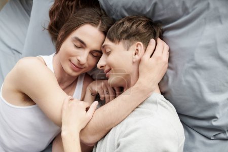 Draufsicht auf lächelndes homosexuelles Paar mit geschlossenen Augen, die sich umarmen und berühren, während sie zusammen auf einem bequemen und gemütlichen Bett zu Hause liegen 