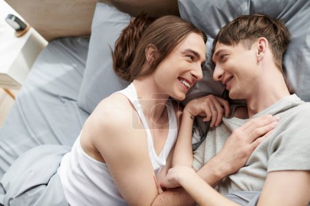 Téléchargez les photos : Vue du dessus du couple homosexuel joyeux et tatoué en vêtements de nuit parlant entre eux tout en étant couchés ensemble sur le lit dans la chambre moderne le matin à la maison - en image libre de droit