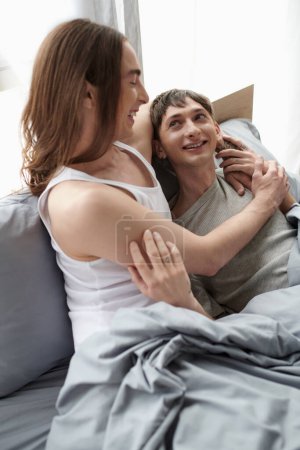 top view of young long haired homosexual man in sleepwear hugging and looking at happy  boyfriend while relaxing together on bed in modern bedroom in morning 