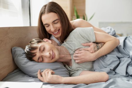 Hombre homosexual despreocupado y de pelo largo abrazando a su novio dormido y sonriente mientras yacen juntos en pijama en una cómoda cama en casa por la mañana 