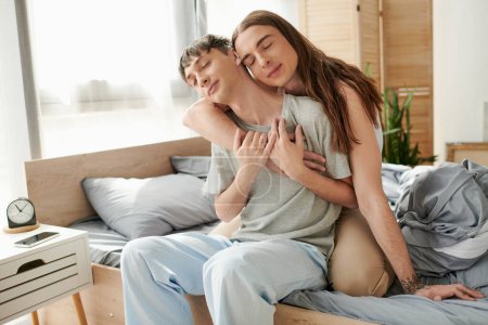 Young long haired homosexual man with closed eyes embracing boyfriend in sleepwear while sitting on bed near cellphone with blank screen and alarm clock on bedside table in morning 