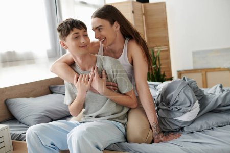 Long haired and tattooed gay man hugging and talking to smiling boyfriend in pajama while sitting together on bed in modern bedroom in morning time 