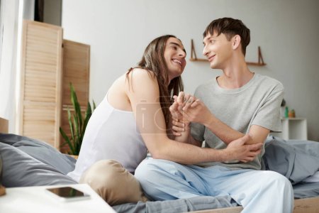 Smiling and young gay couple in pajama and sleepwear holding hands and talking while sitting on comfortable bed near blurred smartphone with blank screen on bedside table at home 