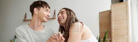 happy same sex couple in sleepwear smiling while holding hands during conversation and looking at each other at modern home in morning time, banner 