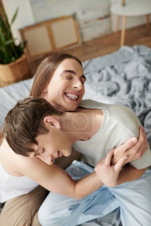 Foto de Vista de ángulo alto de hombre homosexual alegre y de pelo largo en ropa de dormir abrazando novio con los ojos cerrados en la cama borrosa en casa por la mañana - Imagen libre de derechos