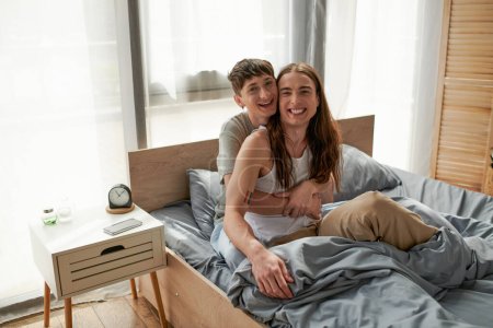 Foto de Joven sonriente pareja del mismo sexo en ropa de dormir abrazando y mirando a la cámara mientras descansa en la cama cerca de teléfono inteligente y despertador en la mesita de noche en casa por la mañana - Imagen libre de derechos