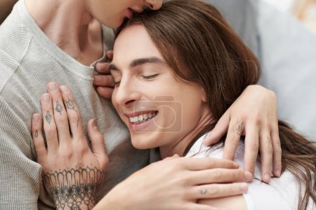Young gay man in sleepwear hugging and kissing head of smiling and tattooed partner with closed eyes on blurred bed at home in morning time 