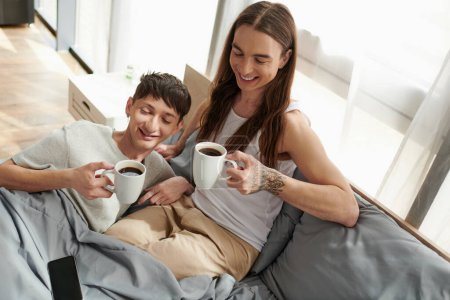High angle view of smiling gay couple in sleepwear holding coffee cups near smartphone with blank screen while resting on comfortable bed at home in morning 