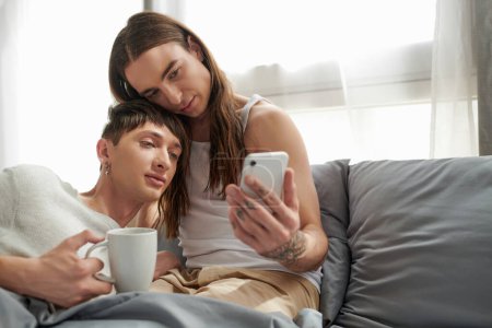 Foto de Joven hombre gay en ropa de dormir sosteniendo la taza de café y mirando el teléfono inteligente mientras está sentado junto con su novio en la cama cómoda en la mañana - Imagen libre de derechos