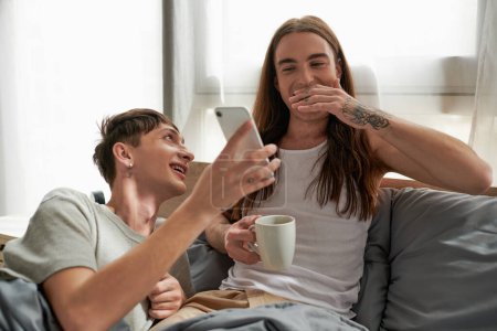 Fröhlicher und junger homosexueller Mann zeigt seinem glücklichen langhaarigen Partner im Pyjama sein Handy, hält eine Kaffeetasse in der Hand und ruht morgens im modernen Schlafzimmer auf dem Bett 