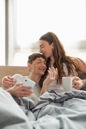 Langhaariger und tätowierter schwuler Mann mit Kaffeetasse und küssendem lächelnden Freund im Pyjama, während er morgens mit Smartphone in der Hand im modernen Schlafzimmer liegt 