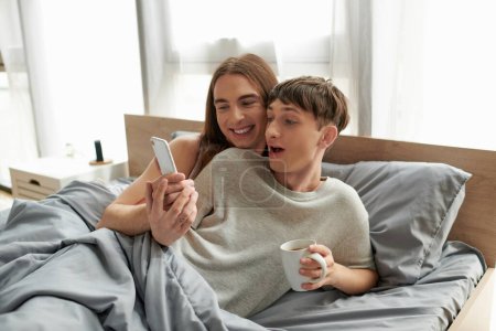 Photo for Smiling long haired gay man showing smartphone at excited boyfriend in pajama holding coffee cup while lying under blanket on bed at home in morning - Royalty Free Image