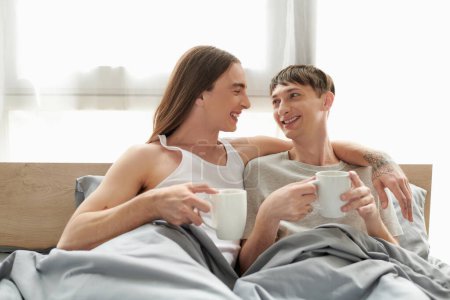 Smiling tattooed gay man in sleepwear hugging and looking at young happy boyfriend while holding coffee cup and resting on comfortable bed in morning at home 