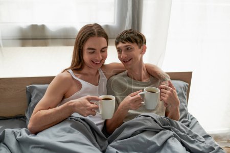 Lächelnder langhaariger und tätowierter schwuler Mann in Nachtwäsche, der eine Kaffeetasse hält und seinen jungen Freund umarmt, während er morgens zu Hause auf einem bequemen Bett aufwacht 