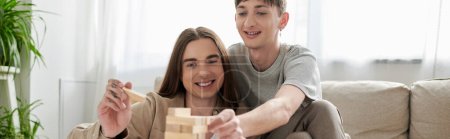 Young and smiling same sex couple in casual clothes playing blurred wood blocks game near couch and blurred green plants in living room at home, banner 
