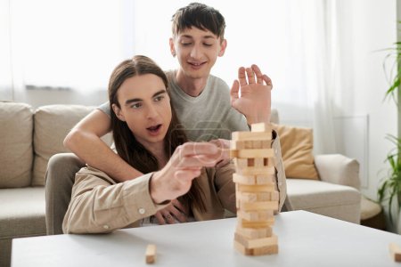 Young and positive gay man hugging long haired boyfriend in casual clothes while playing blurred wood blocks game on table near comfortable couch in living room at home 