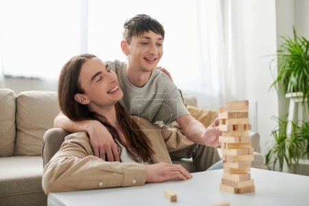 Hombre homosexual despreocupado en ropa casual abrazando novio de pelo largo y jugando bloques de madera juego en la mesa mientras pasan tiempo juntos en la sala de estar en casa 