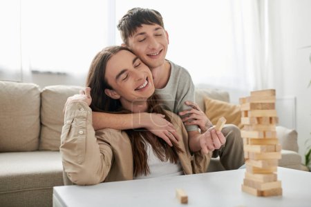 Junge lächelnde Homosexuelle mit geschlossenen Augen umarmen langhaarigen Freund in lässiger Kleidung in der Nähe verschwommenen Holzblöcke Spiel auf dem Tisch im Wohnzimmer zu Hause 