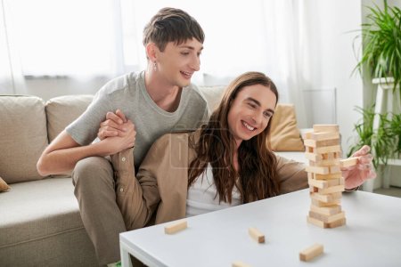 Young and smiling same sex couple in casual clothes holding hands while playing wood blocks game near parts on table in living room at home 