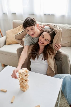 Positive and long haired homosexual man hugging boyfriend while playing wood blocks game on table near comfortable couch in living room at home 