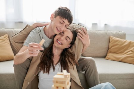 Despreocupado joven pareja homosexual en ropa casual cerrando los ojos mientras abraza y juega bloques de madera borrosa juego cerca del sofá en la sala de estar en casa 