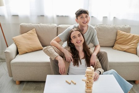 Lächelndes und junges homosexuelles Paar in lässiger Kleidung, das in der Nähe von verschwommenen Holzblöcken auf dem Tisch in die Kamera schaut, während es zu Hause auf der Couch im Wohnzimmer sitzt 