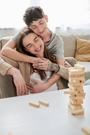 Smiling gay man with closed eyes hugging tattooed and long haired boyfriend in casual clothes near blurred wood blocks game and parts on table in living room at home  