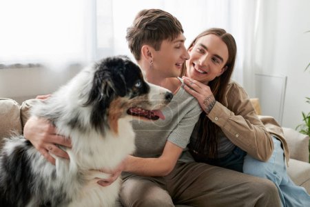 Cheerful and long haired gay man with tattoo on hand hugging and looking at young boyfriend near blurred Australian shepherd dog sitting on couch in living room at home 