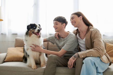 Überglücklich langhaariger schwuler Mann in lässiger Kleidung umarmt Freund streichelt pelzigen australischen Schäferhund, während er auf bequemer Couch im Wohnzimmer zu Hause sitzt 
