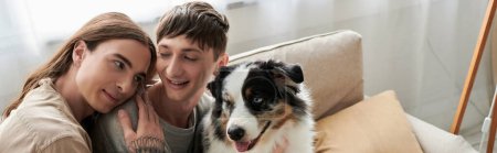 Long haired gay man with tattoo on hand hugging smiling boyfriend in casual clothes near Australian shepherd dog on modern couch in living room at home, banner 