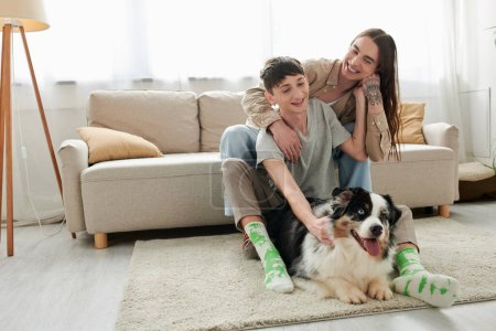 Cheerful tattooed gay man hugging young boyfriend in casual clothes and socks while looking at Australian shepherd dog lying on carpet near couch in modern living room at home  