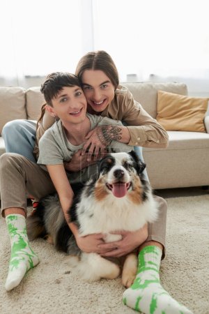 Alegre y joven pareja lgbt en ropa casual y calcetines abrazando y mirando a la cámara cerca peludo perro pastor australiano acostado en la alfombra en el suelo en la sala de estar moderna en casa 