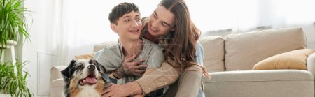 Photo for Tattooed and long haired homosexual man hugging young smiling boyfriend and petting furry Australian shepherd dog near couch and green plants in modern living room at home, banner - Royalty Free Image