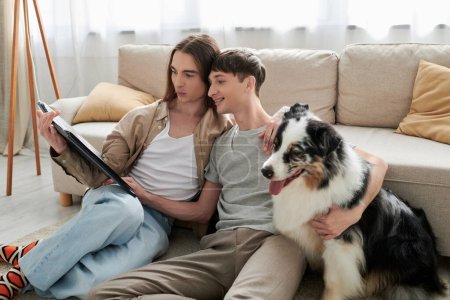 Lächelndes schwules Paar in lässiger Kleidung und Socken beim Betrachten eines geöffneten Fotoalbums neben pelzigem australischen Schäferhund und bequemer Couch im heimischen Wohnzimmer 
