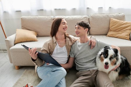 Photo for Carefree homosexual couple in casual clothes talking while spending time with Australian shepherd dog and holding photo album in modern living room at home - Royalty Free Image