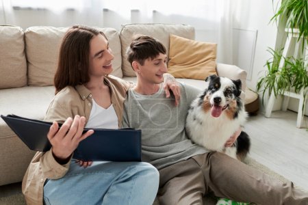 Alegre y joven pareja del mismo sexo abrazándose mientras sostiene álbum de fotos y mirando perro pastor australiano amigable en el suelo en la sala de estar moderna en casa 