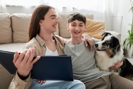 Positive same sex couple in casual clothes looking at blurred photo album and hugging friendly Australian shepherd dog near couch in living room at home 