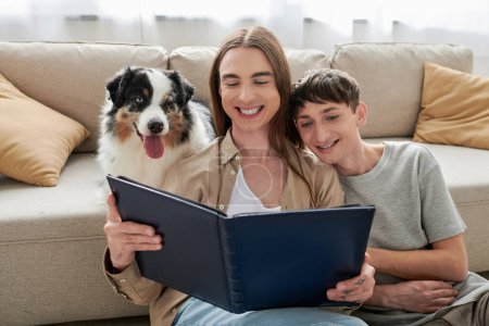 Überglückliches lgbt-Paar schaut auf Fotoalbum, während es neben australischem Schäferhund und gemütlicher Couch im modernen Wohnzimmer zu Hause sitzt 