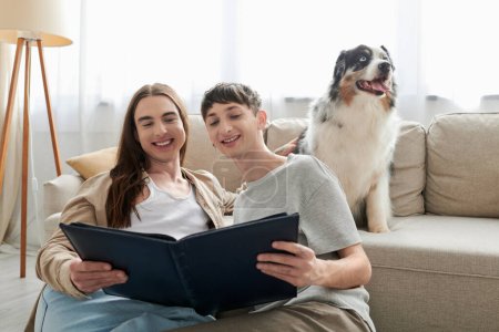 Positives lgbt Paar schaut auf Fotoalbum und lächelt, während es fröhliche Erinnerungen hat und neben australischem Schäferhund und Couch im modernen Wohnzimmer zu Hause sitzt 