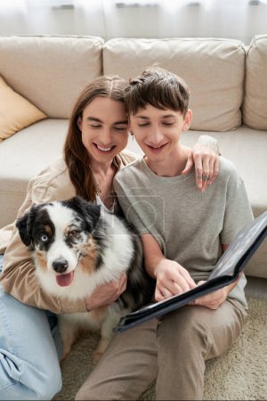Photo for Happy gay couple looking together at photo album and smiling while having happy memories and sitting near Australian shepherd dog and sofa in modern living room - Royalty Free Image