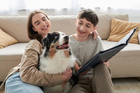 smiling and young gay men looking together at Australian shepherd dog and holding photo album while smiling in living room at modern apartment 
