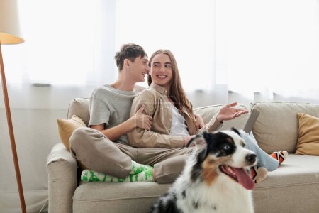 happy gay man with long hair pointing with hand at laptop while sitting on couch with happy boyfriend in casual clothes near Australian shepherd dog inside of living room in modern apartment 