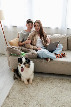 happy lgbt couple in casual clothes sitting on couch with laptop and smiling while looking at Australian shepherd dog on carpet inside of living room in modern apartment 
